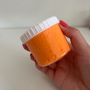 Peut inclure: Un récipient cylindrique en plastique transparent rempli d'une substance texturée orange vif. Le récipient est doté d'un couvercle blanc à visser avec des rainures. La matière orange à l'intérieur présente un aspect poreux et irrégulier.