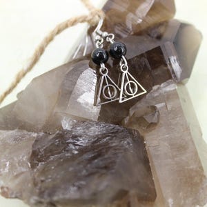May include: Silver earrings with black beads and a triangle charm with the Deathly Hallows symbol. The earrings are displayed on a cluster of brown crystals.