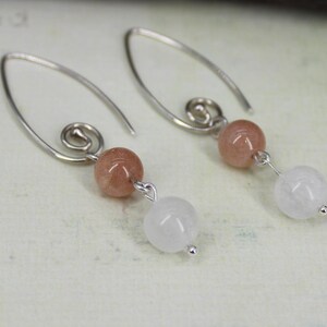 May include: Silver dangle earrings with a peach-colored gemstone and a white gemstone. The earrings have a spiral design at the top.