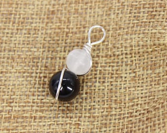 May include: A pendant featuring a black and a white gemstone, wire-wrapped in silver. The pendant has a loop at the top for hanging. The gemstones are round and smooth. The background is a woven, tan fabric.