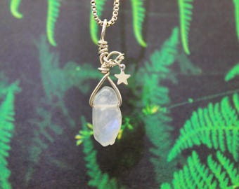 May include: A silver-toned necklace featuring a raw, pale blue moonstone pendant. The pendant is wire-wrapped and accented with a small star charm. The chain is a delicate box chain. The background is a blurred image of green foliage.