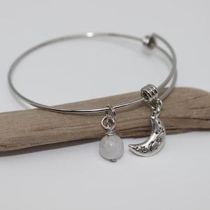 May include: A silver-toned wire bangle bracelet featuring two dangling charms. One charm is a smooth, round, pale white stone bead, and the other is a silver-toned crescent moon charm decorated with small star cutouts along its curved surface.
