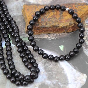 May include: A black beaded necklace and bracelet set. The necklace is long and draped, while the bracelet is a smaller, circular design. Both pieces feature dark, glossy beads, and are displayed on a natural stone.