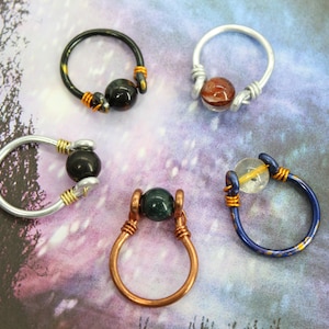 May include: Five wire wrapped rings with different colored wire and gemstone beads. The rings are on a purple and blue background.