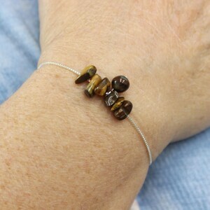 May include: A delicate bracelet featuring a string of small, polished tiger's eye stones in shades of brown and gold. The stones are strung on a thin, silver-colored chain. The bracelet is worn on a wrist.