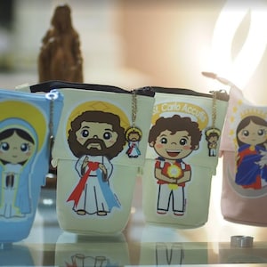 Könnte beinhalten: Vier farbenfrohe Reißverschlusstaschen mit Bildern von religiösen Figuren. Jede Tasche hat eine goldene Kette mit einem kleinen Anhänger. Die Taschen befinden sich auf einer Glasoberfläche.
