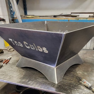 May include: A large, square, metal fire pit with a raised base. The fire pit is made of dark metal and has the words "The Coles" cut out of the front.