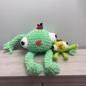 May include: Two handmade crochet monster toys. The larger monster is light green with white eyes, a red patch, and a colorful headpiece. The smaller monster is lime green with similar features. Both are on a wooden surface.