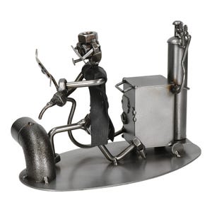 Puede incluir: Escultura metálica de un soldador en el trabajo. La figura está hecha de tuercas, pernos y varillas de metal, con un delantal negro. Un tanque de soldadura y equipo están cerca, todo sobre una base de metal. Una pieza única de arte industrial.