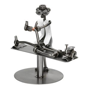 May include: A metal sculpture depicting a doctor and patient. The doctor figure, made of nuts and bolts, wears a white coat and holds a medical instrument. The patient lies on a metal table. The sculpture is mounted on a round base.