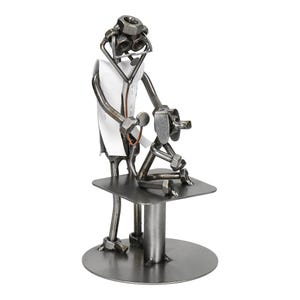 Puede incluir: Escultura metálica que representa a un veterinario examinando a un perro. El veterinario, hecho de tuercas y tornillos, lleva una bata blanca y usa un estetoscopio. El perro está sentado en una mesa de metal. La escultura está sobre una base redonda.