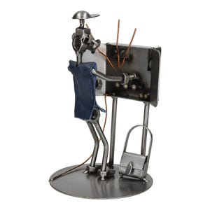 Könnte beinhalten: Eine Metallskulptur einer schweißenden Person, hergestellt aus Muttern, Schrauben und Metallteilen. Die Figur trägt eine blaue Lederschürze und eine Kappe. Eine kleine Handtasche steht daneben. Die Skulptur ist auf einer kreisförmigen Basis montiert.