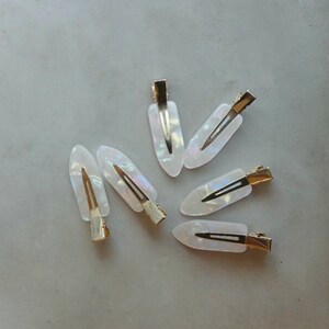 May include: Six white iridescent hair clips with gold metal clasps. The clips are shaped like a teardrop and have a smooth, glossy finish.