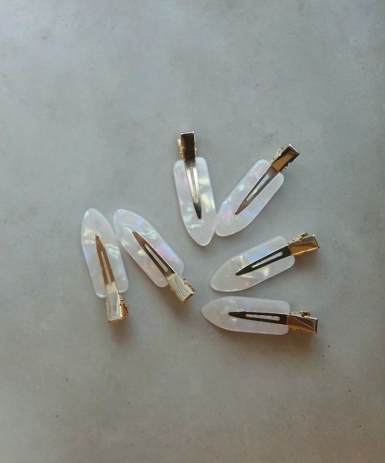 May include: Six white iridescent hair clips with gold metal clasps. The clips are shaped like a teardrop and have a smooth, glossy finish.