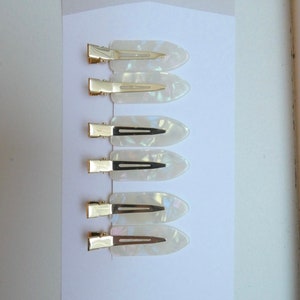 May include: Set of six white, pearlescent hair clips with gold metal clasps. The clips are arranged vertically on a white card with the text "JILL BUCK HAIR" and "CREASELESS HAIR CLIPS".