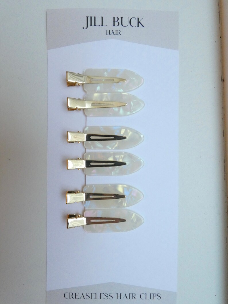 May include: Set of six white, pearlescent hair clips with gold metal clasps. The clips are arranged vertically on a white card with the text "JILL BUCK HAIR" and "CREASELESS HAIR CLIPS".
