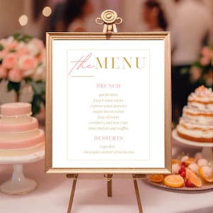 May include: A framed menu sign with gold lettering and a pink accent, displayed on a gold easel. The menu lists brunch and dessert items, including quiche bites, fruit skewers, and cupcakes. The sign is set up at an event with blurred background elements.