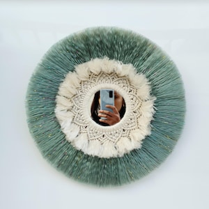 Peut inclure: Un miroir rond en macramé avec un centre blanc et un anneau extérieur vert. Le miroir est décoré de franges et de glands.