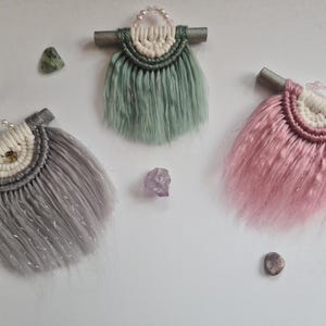 Peut inclure: Trois petites tentures murales avec des franges, chacune d'une couleur différente : vert, rose et argent. Les tentures sont faites de fil et ont un bâtonnet de bois en haut.