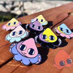 Pode incluir: Uma coleção de alfinetes de esmalte coloridos com um design de polvo de desenho animado. Os alfinetes têm uma variedade de cores, incluindo rosa, amarelo, roxo e azul, com contornos pretos. A palavra "Squiddy" está escrita em azul claro no topo.
