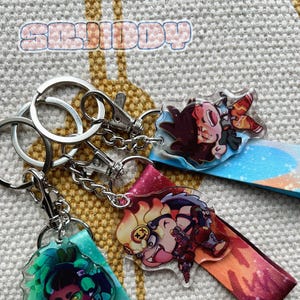 May include: Four colourful keychains featuring different cartoon characters. Each keychain has a clear acrylic charm with a colourful background and a silver metal ring and chain.