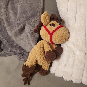 Könnte beinhalten: Ein handgefertigtes, gehäkeltes Pferdespielzeug in Beige- und Brauntönen mit rotem Halfter. Das Spielzeug hat eine flauschige Textur und liegt auf einer grauen Oberfläche neben einer weißen, strukturierten Decke. Das Pferd hat dunkelbraune Hufe und einen Schwanz.