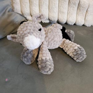 Könnte beinhalten: Ein handgefertigtes Kuscheltier in Form eines Häschens. Das Häschen ist überwiegend grau mit weißen Akzenten im Gesicht und schwarzen Details an den Ohren. Das Stofftier liegt auf einer grauen Oberfläche.