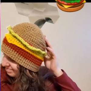 May include: A brown crocheted hat shaped like a hamburger with yellow, green, and red details. The hat is being worn by a person.