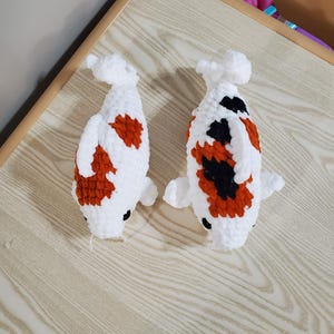Könnte beinhalten: Zwei handgefertigte Koi-Fische aus Häkelarbeit. Die Fische sind weiß mit orangefarbenen und schwarzen Flecken und haben kleine schwarze Augen. Die Koi liegen auf einer hellen Holzmaserung.
