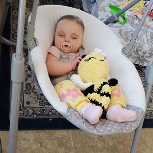 Può includere: Un bambino in un'altalena per bambini bianca e grigia con un giocattolo a forma di ape in crochet giallo e nero. Il bambino indossa un vestito rosa e giallo con fiori rosa e calzini bianchi.