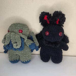 Op de afbeelding: Twee handgemaakte gehaakte knuffels. De ene is een saliegroen wezen met een tentakelgezicht, blauwe accenten en rode ogen. De andere is een zwart pluizig wezen met grote oren, rode details aan de binnenkant van de oren en rode ogen.