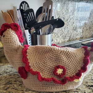 May include: A crocheted brown chicken pot holder with red and white accents. The chicken has a yellow beak and a red comb.