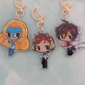 Puede incluir: Tres llaveros acrílicos con herrajes dorados. Cada llavero presenta un diseño de personaje de dibujos animados. Los personajes tienen diferentes colores de cabello y visten varios atuendos. Los llaveros están sobre una superficie azul claro y blanca.