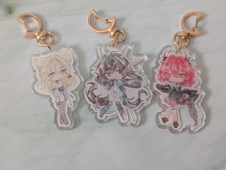 Pode incluir: Tr&ecirc;s chaveiros de acr&iacute;lico em estilo anime com fechos dourados. Cada chaveiro apresenta uma ilustra&ccedil;&atilde;o de personagem diferente em tons de branco, preto, rosa e azul. Os chaveiros s&atilde;o cortados em forma dos personagens.