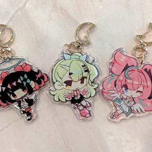 May include: Three acrylic keychains featuring anime-style characters with gold-colored crescent moon clasps. The characters have colorful hair and outfits, with one in black and pink, one in green, and one in pink and blue. Each keychain is a unique design.