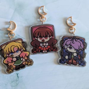 Op de afbeelding: Drie karaktercharms met gouden sleutelhangers. Elke charme heeft een ander anime-personage met een bloemenontwerp. De personages zijn een meisje met bruin haar, een meisje met blond haar en een jongen met paars haar.