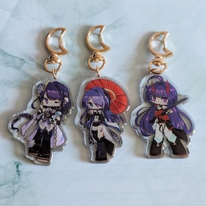 May include: Three acrylic keychains featuring anime-style characters with purple and black outfits. Each keychain has a gold-colored crescent moon clasp. One character holds a red umbrella. The keychains are on a light blue and white surface.