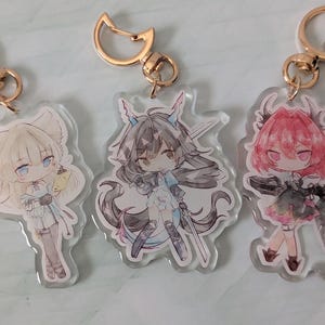 Pode incluir: Três chaveiros de acrílico em estilo anime com acessórios dourados. Cada chaveiro apresenta um personagem diferente com cabelo e roupas exclusivos. Os personagens são em tons de branco, preto e rosa.