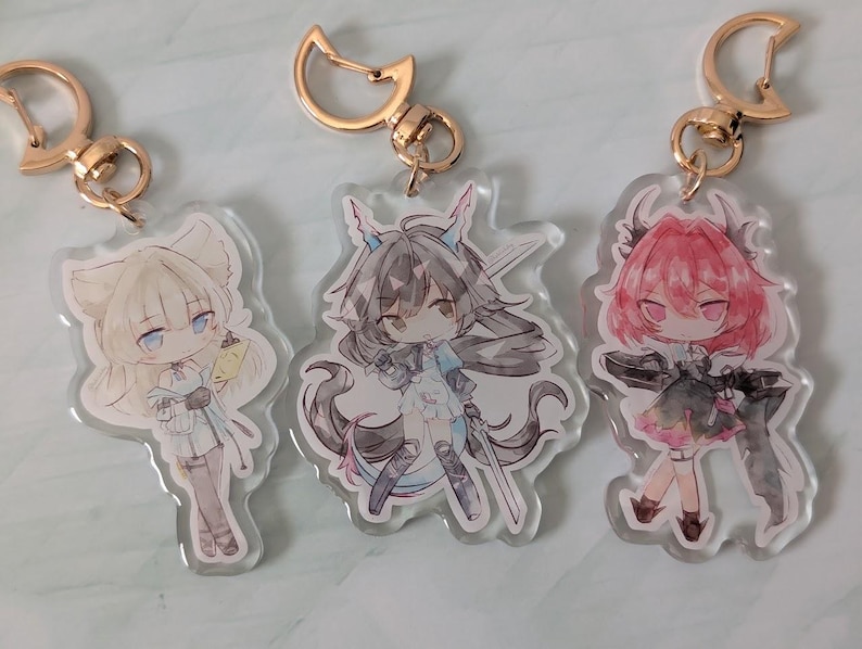 Pode incluir: Tr&ecirc;s chaveiros de acr&iacute;lico em estilo anime com acess&oacute;rios dourados. Cada chaveiro apresenta um personagem diferente com cabelo e roupas exclusivos. Os personagens s&atilde;o em tons de branco, preto e rosa.