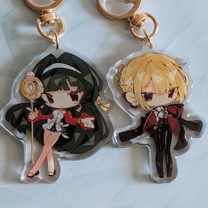 May include: Two anime character keychains with gold chains. The keychain on the left features a girl with long black hair, a red and white dress, and a gold wand. The keychain on the right features a girl with long blonde hair, a red and black coat, and black pants.
