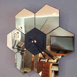 Op de afbeelding: Een zeshoekige spiegelklok met zwarte wijzers en een zwart midden. De klok bestaat uit zes gespiegelde zeshoeken, die elk verschillende delen van een kamer reflecteren.