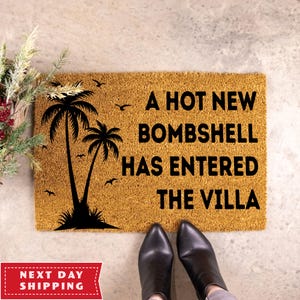 May include: A rectangular tan coir doormat featuring a black graphic of two palm trees and several birds. The text A HOT NEW BOMBSHELL HAS ENTERED THE VILLA is printed in bold black capital letters on the right side of the mat surface.