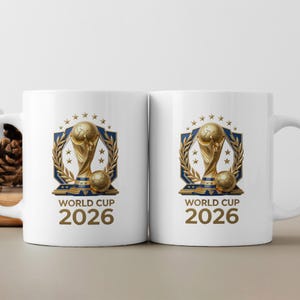 May include: Two white ceramic mugs featuring a gold-colored trophy graphic centered on the front. The design includes a blue shield, golden laurel branches, stars, and a soccer ball. Below the graphic, the text reads WORLD CUP 2026 in gold letters.