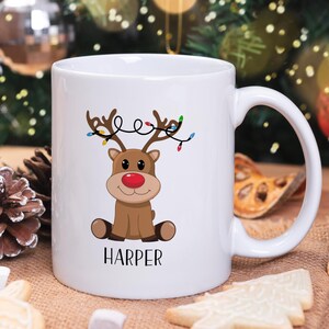 May include: White ceramic mug with a cartoon reindeer wearing a string of Christmas lights. The reindeer is brown with a red nose and is sitting down. The mug has the name "HARPER" printed on the bottom.