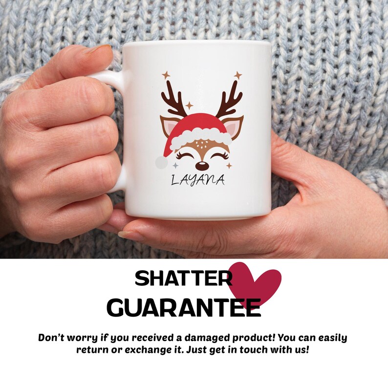 May include: White ceramic mug with a red and brown reindeer illustration wearing a Santa hat. The mug has the name "LAYANA" printed on it. The mug is being held by a person's hand.
