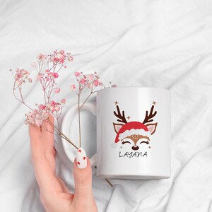 May include: White ceramic mug with a cartoon reindeer wearing a Santa hat. The reindeer has closed eyes and a smile. The mug has the name "Layana" printed on it.