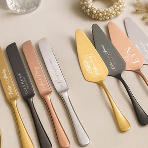 May include: An assortment of personalized cake servers and butter knives in gold, black, silver, and rose gold. Each piece is engraved with names and dates, perfect for weddings or special events. The utensils are arranged on a light-colored surface.