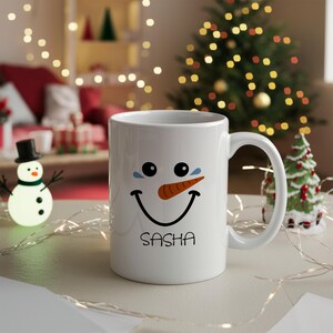 May include: White ceramic mug with a snowman face and the name Sasha printed on the bottom. The snowman has a carrot nose and blue eyes.