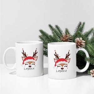 May include: Two white ceramic mugs with a reindeer design wearing a Santa hat. The mugs have the name "Layana" printed on them.