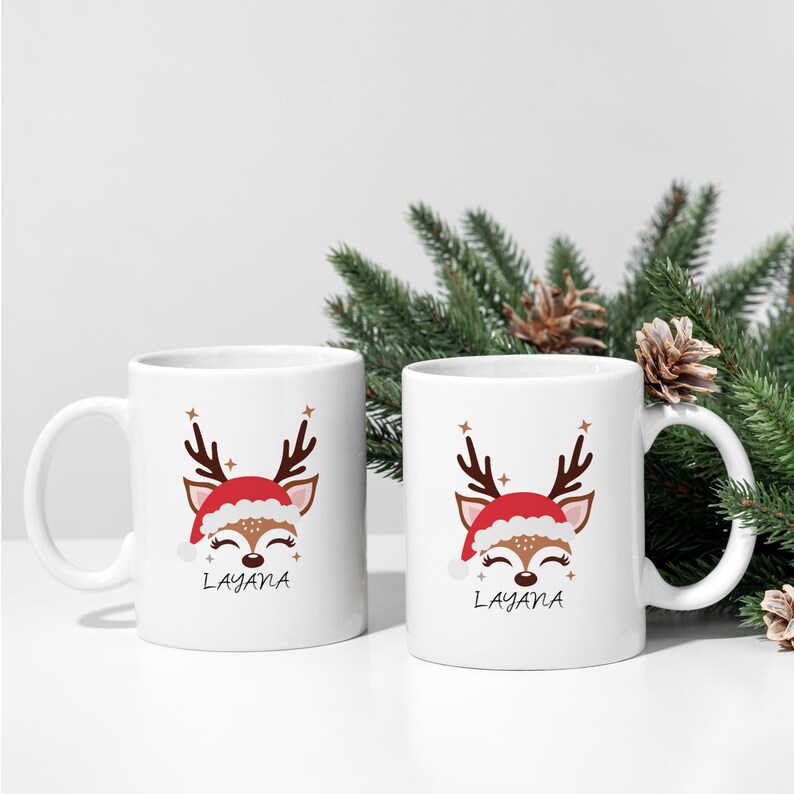 May include: Two white ceramic mugs with a reindeer design wearing a Santa hat. The mugs have the name "Layana" printed on them.